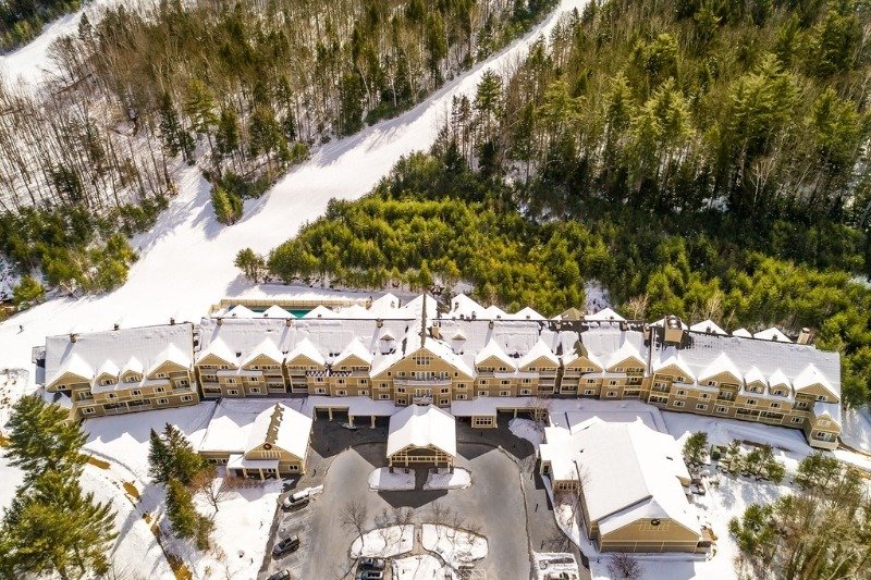 Aerial winter view of Grand Summit Hotel and Attitash Mountain Resort in Bartlett, New Hampshire showing the snow-covered resort buildings, surrounding forest, ski trails, and scenic White Mountains landscape.