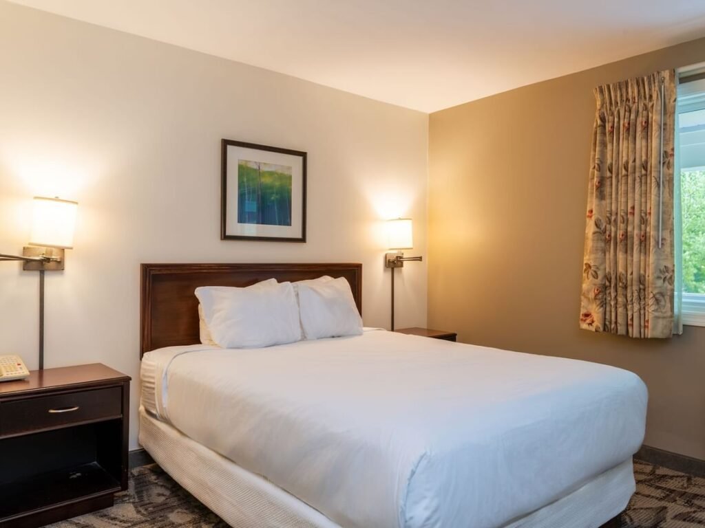 Cozy bedroom inside Unit 335 Interval 1 at Grand Summit Hotel in Bartlett, New Hampshire featuring a queen bed with white linens, bedside tables, warm lighting, neutral walls, and a window with patterned curtains in this inviting White Mountains retreat.